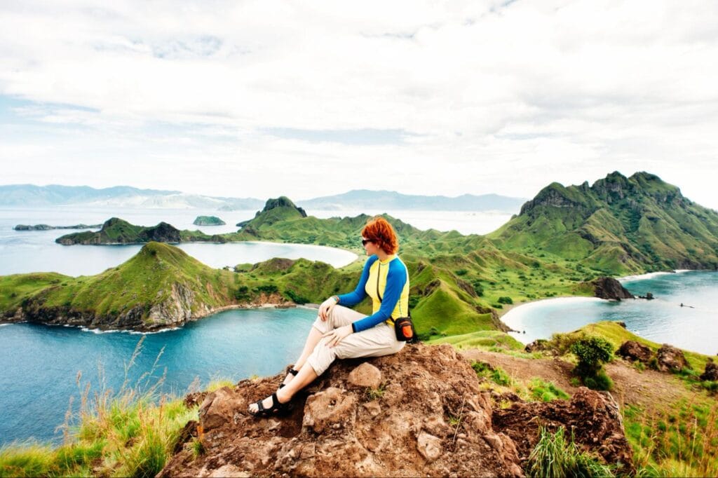 solo traveler duduk menikmati pemandangan pulau padar dengan bukit hijau dan laut biru dari atas